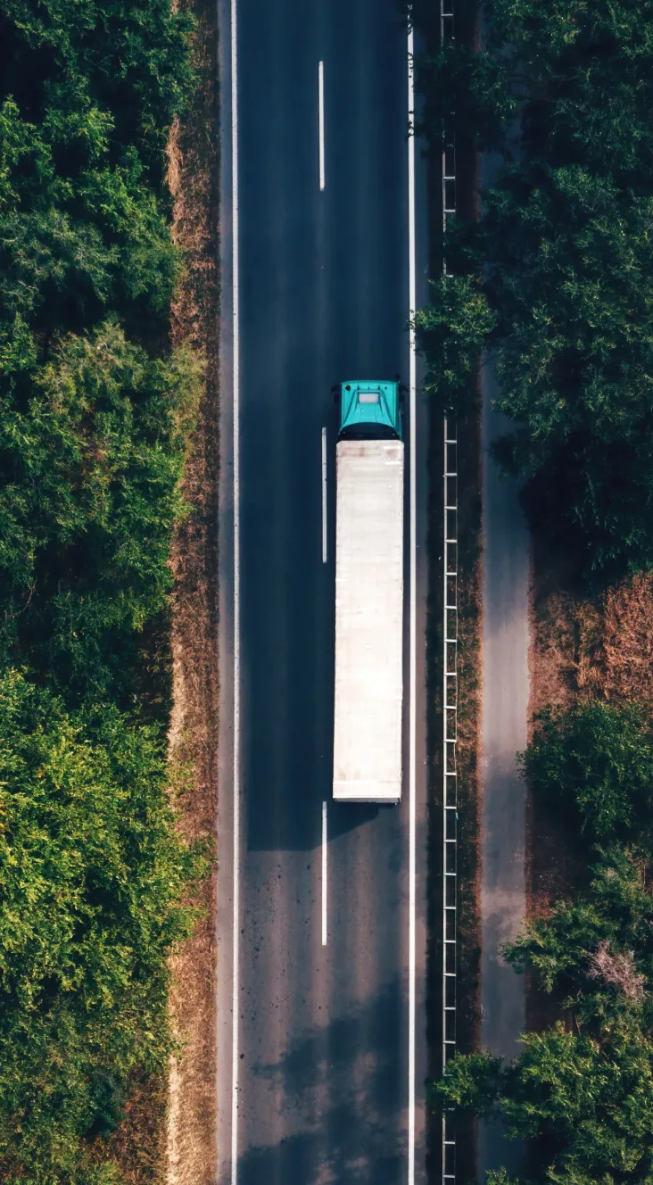 Aerial view of a teal semi-truck with a white trailer driving on a two-lane highway surrounded by green trees.
