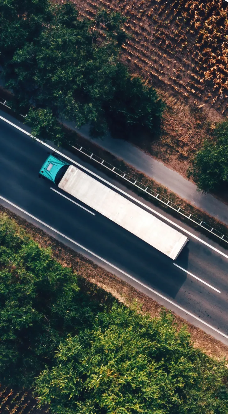 Aerial view of a teal semi-truck with a white trailer driving on a two-lane highway surrounded by green trees.