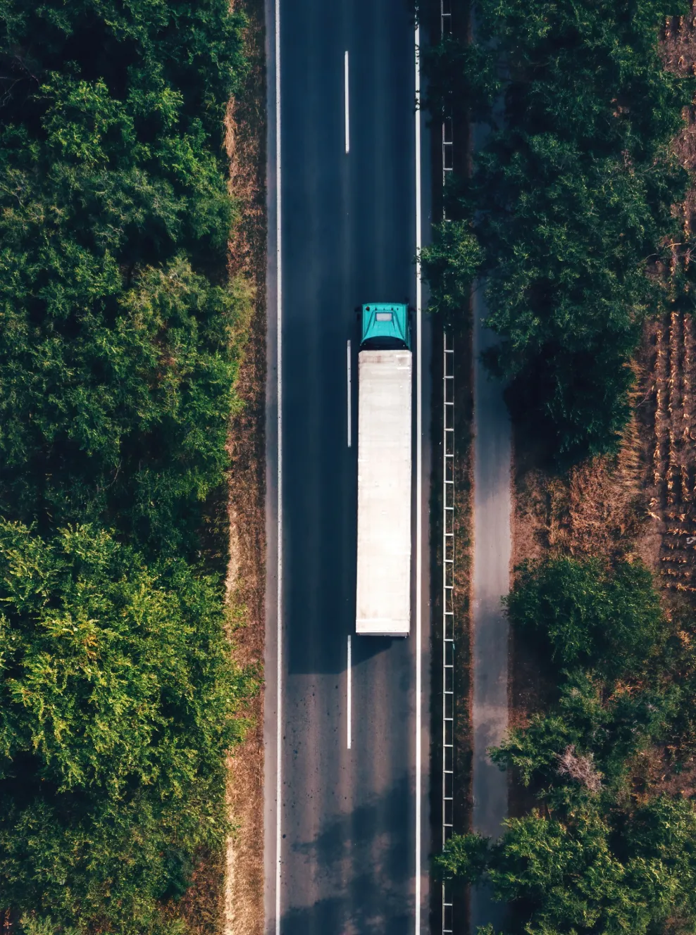 Aerial view of a teal semi-truck with a white trailer driving on a two-lane highway surrounded by green trees.